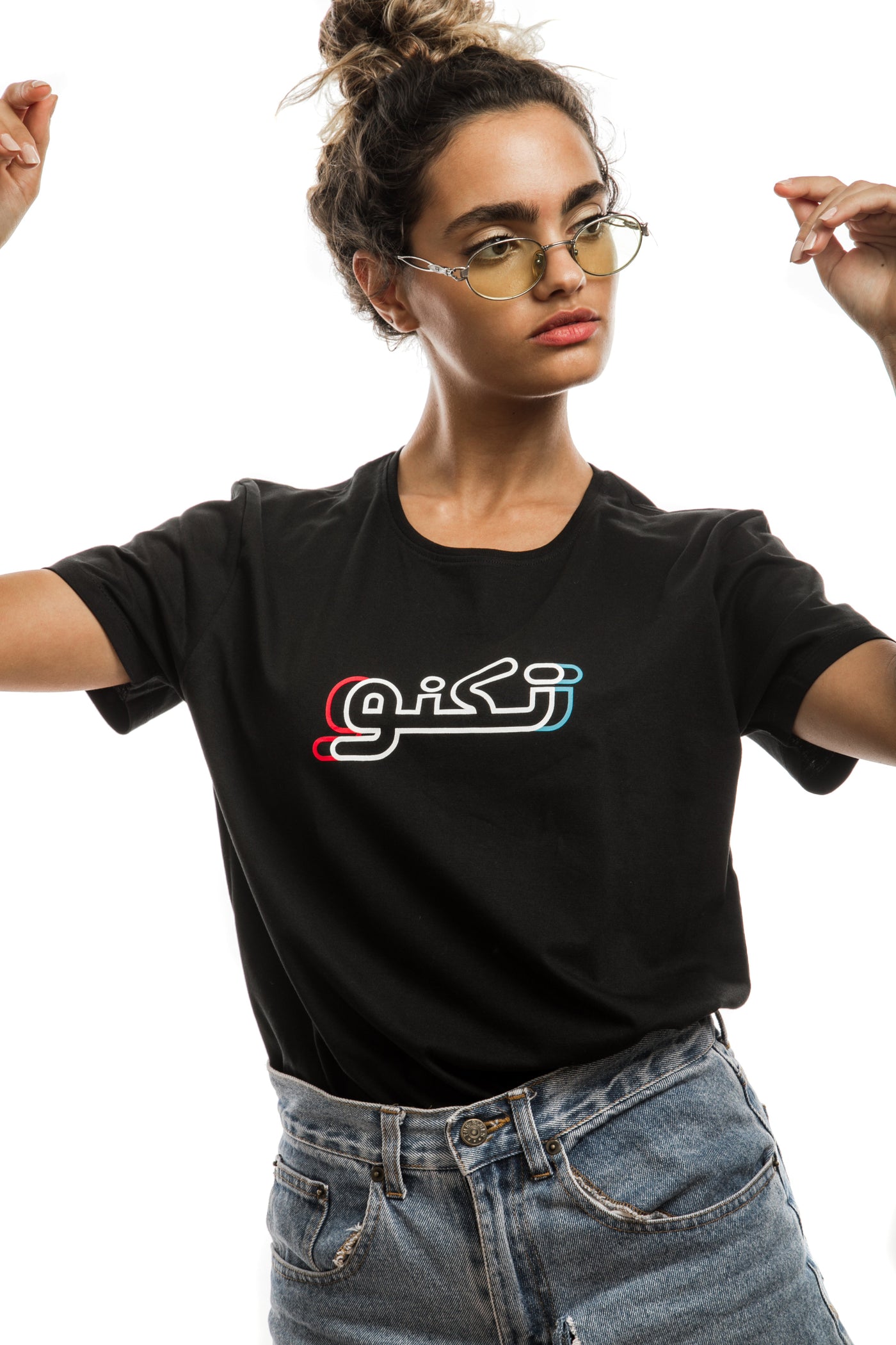 young adult female wearing black t-shirt with techno written in arabic تكنو and in blue, red and white in the center of the t-shirt along with blue jeans shorts and glasses