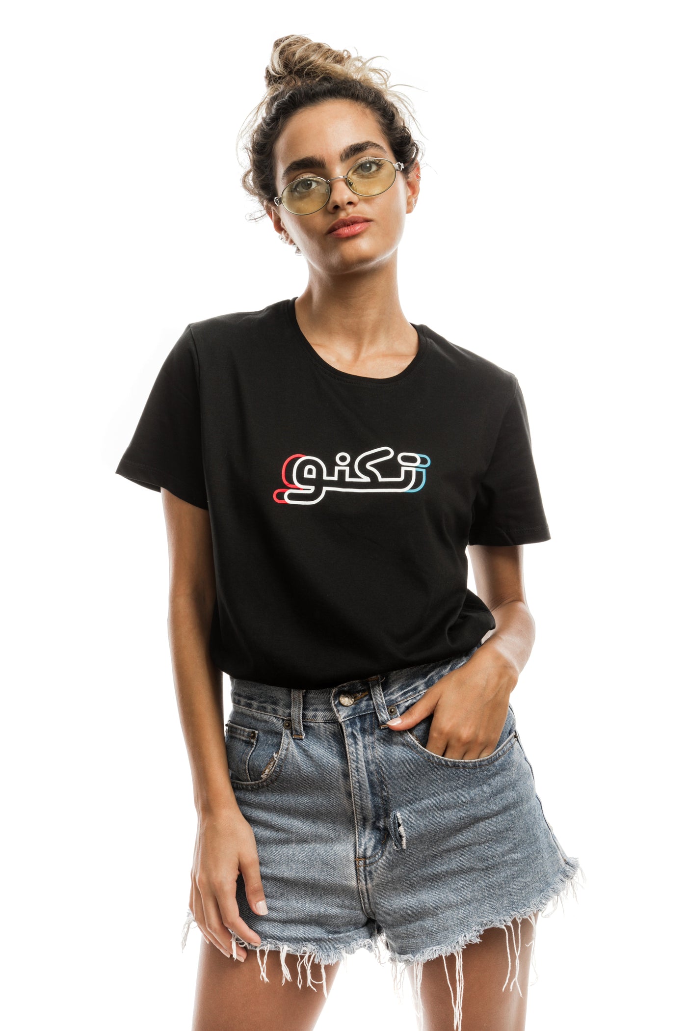 young adult female wearing black t-shirt with techno written in arabic تكنو and in blue, red and white in the center of the t-shirt along with blue jeans shorts and glasses
