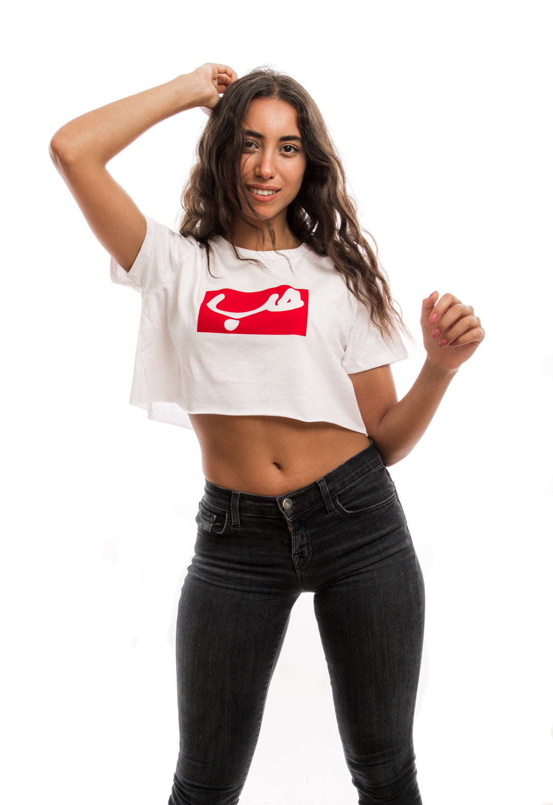 young adult female wearing white crop top with hobb written in arabic حب in white and in a red frame in the center of the crop top along with dark blue jeans