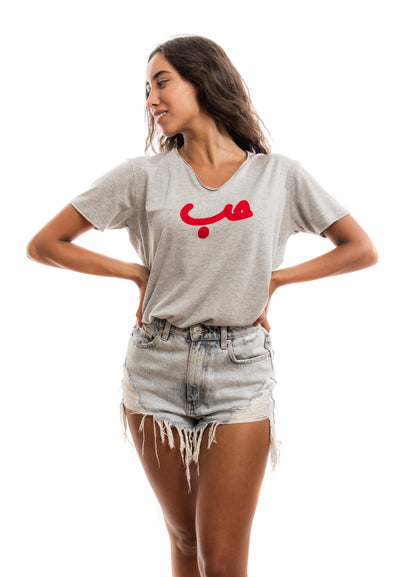 young adult female wearing grey t-shirt with Hobb written in arabic حب and in red velvet in the center of the t-shirt along with light blue jeans shorts
