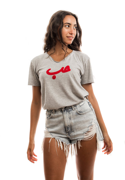 young adult female wearing grey t-shirt with Hobb written in arabic حب and in red velvet in the center of the t-shirt along with light blue jeans shorts