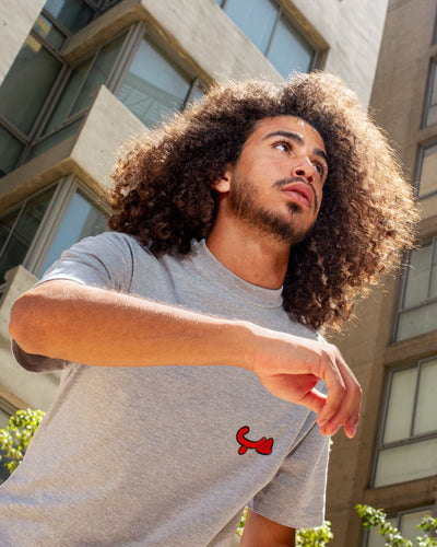 Young adult male wearing an oversized grey t-shirt with Hobb written in arabic حب and in red silkscreen on the side of the shirt. Standing in front of a modern building