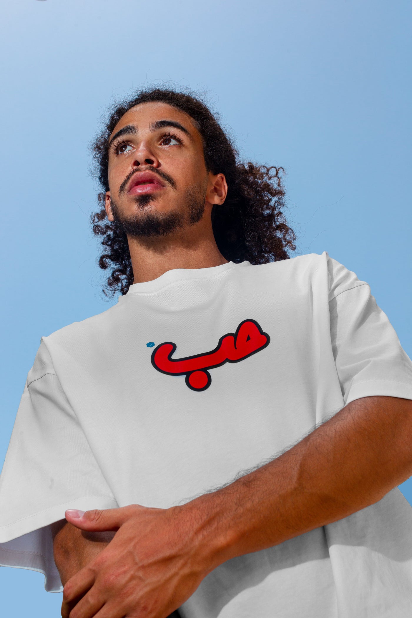 Young adult male wearing an oversized white t-shirt with Verified Hobb written in arabic حب and in red silkscreen in the center of the shirt.