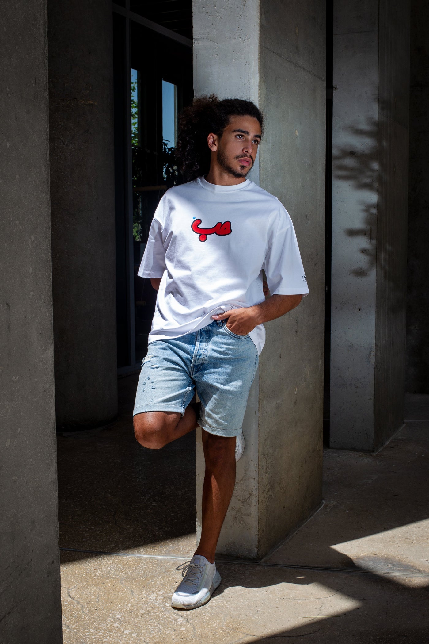 Young adult male wearing an oversized white t-shirt with Verified Hobb written in arabic حب and in red silkscreen in the center of the shirt along with jeans shorts and white sneakers.
