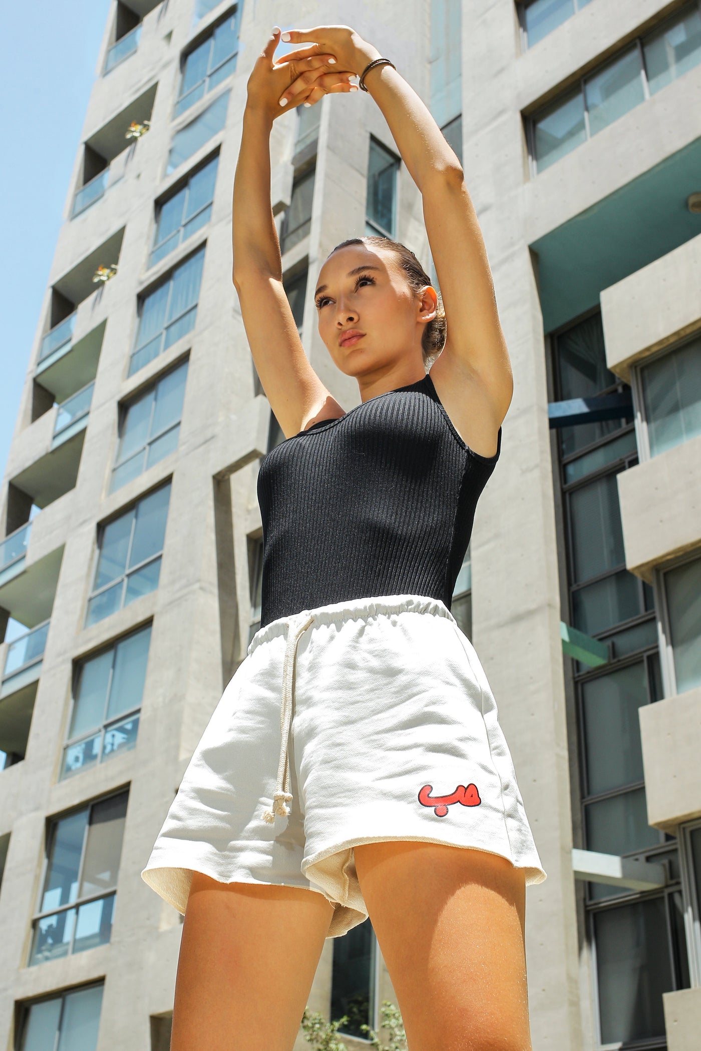 Young adult female wearing off white flared short with Hobb written in arabic حب and in red silkscreen on the side of the short along with a black sleeveless top. Standing in front of a modern building.