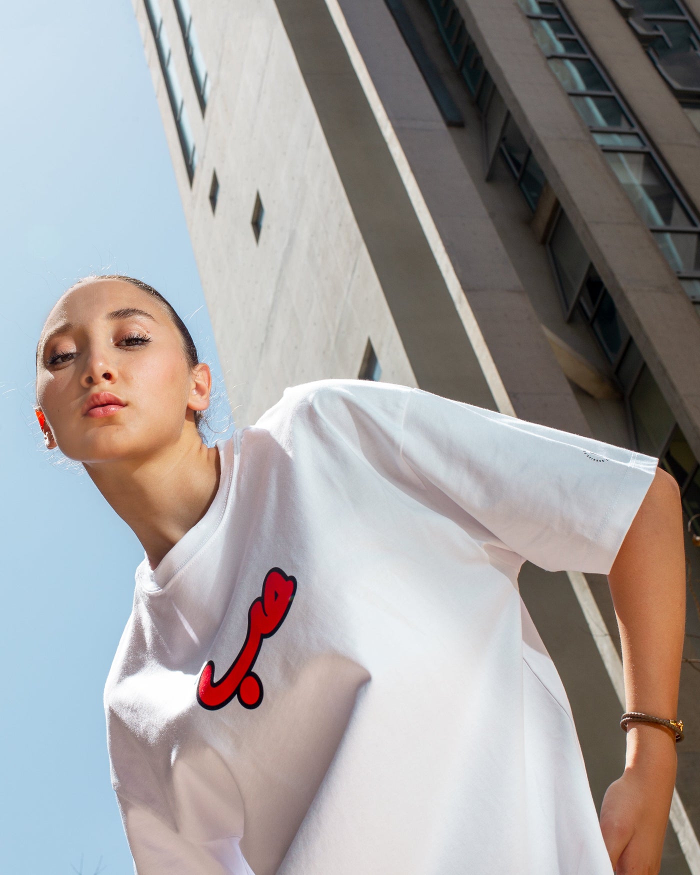 Young adult female wearing an oversized white t-shirt with Verified Hobb written in arabic حب and in red silkscreen in the center of the shirt. Standing in front of a modern building