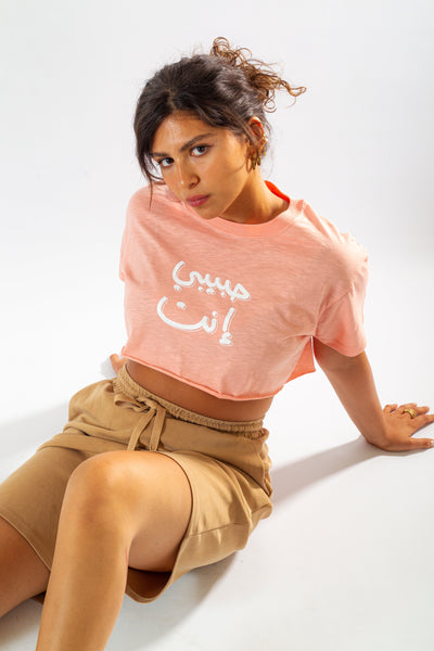 Young adult female wearing an oversized peach crop top with Habibi Enta written in arabic حبيبي إنت and in white silkscreen in the center of the top along with beige shorts.