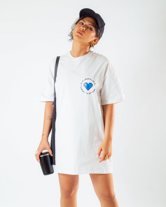 Young adult female wearing an oversized white T-Dress with Khanat Al Sadaka written in arabic خانة الصداقة and in blue and black silkscreen on the side of the dress along with black cap on her head, a black tote bag and a black coffee cup.