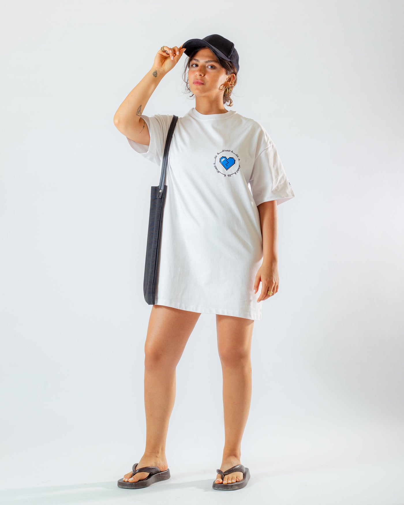 Young adult female wearing an oversized white T-Dress with Khanat Al Sadaka written in arabic خانة الصداقة and in blue and black silkscreen on the side of the dress along with black cap on her head, a black tote bag.
